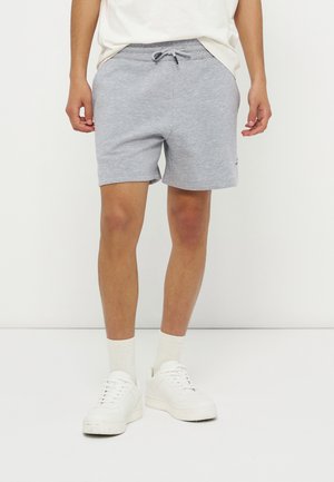 Adulte portant un short gris clair avec un cordon, des chaussettes blanches et des baskets blanches, debout sur un fond blanc uni.