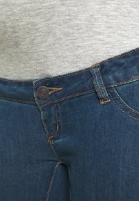Mörka denimjeans med knäppning, orange sömmar och en kroppsnära design. Synliga detaljer av midjeband och fickor.