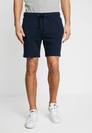 Mann trägt marineblaue Shorts mit Kordelzug, graues T-Shirt und weiße Turnschuhe, steht vor einem schlichten weißen Hintergrund.