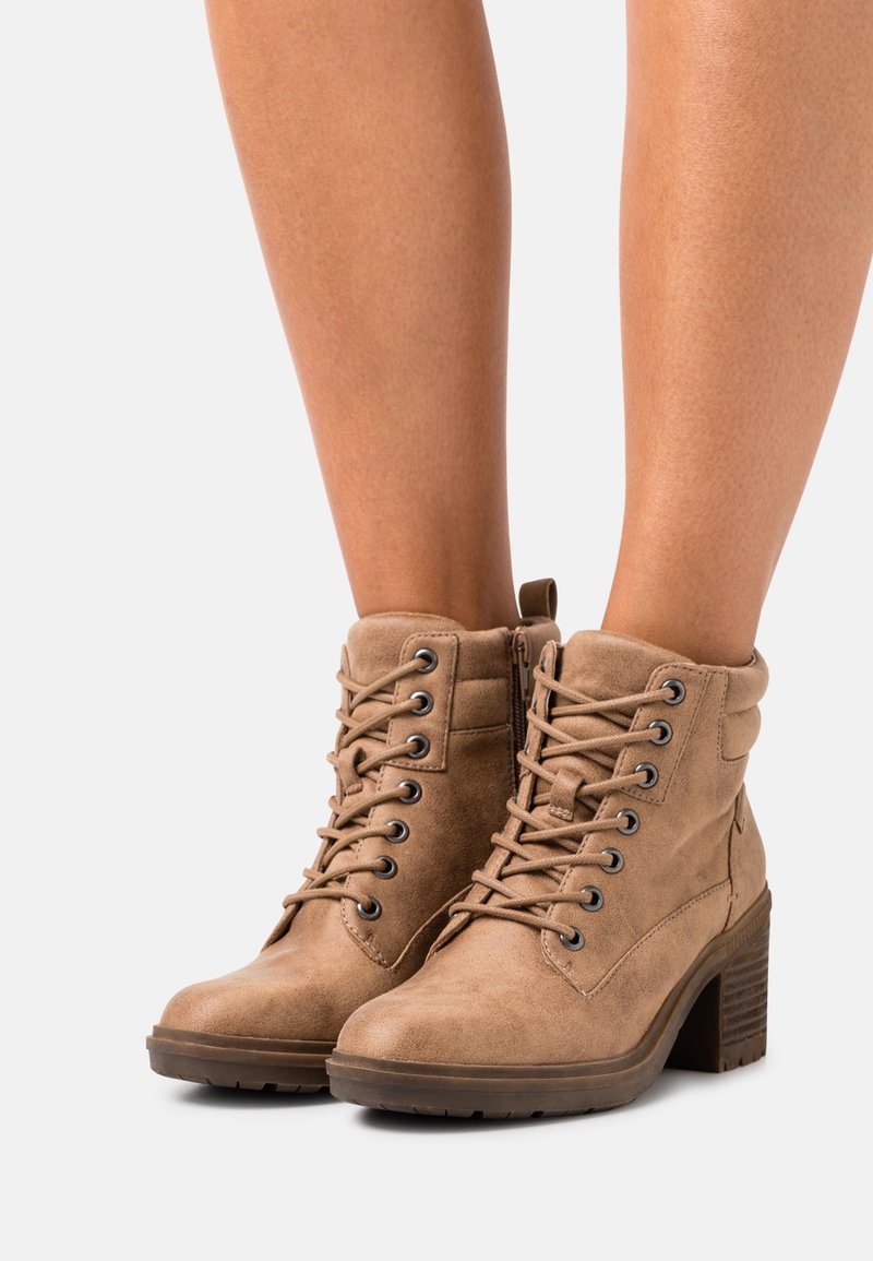 Bottines en daim beige à lacets sur le devant et fermeture éclair latérale, présentant une finition texturée et un talon épais. La quincaillerie comprend des œillets ronds.