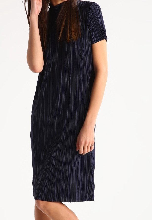 Femme portant une robe plissée bleu foncé à manches courtes et longueur genou, sur un fond blanc uni.