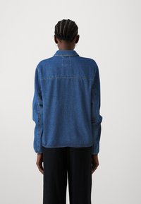 Chaqueta de mezclilla azul con cuello, mangas largas, detalle en el yugo trasero y acentos de costura. Llevada con pantalones negros.