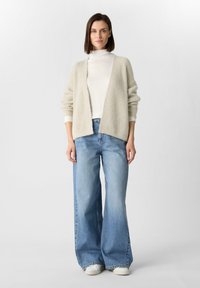 Beige gebreide cardigan met open voorkant, gecombineerd met losse lichtblauwe jeans en witte sneakers. Textuurstof met ribbelmanchetten.