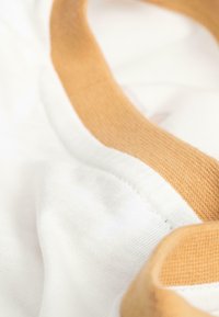 Baumwollstoff mit einem weißen Hauptteil und einem glatten, ockerfarbenen Kragen. Sichtbare Nähte umranden den Kragen und die Stoffstruktur.