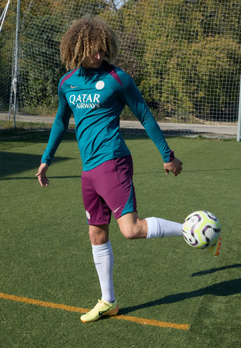 Voetballer in een teal en maroon shirt, met witte sokken, die een trap uitvoert met een gemarmerde bal op een groen veld.