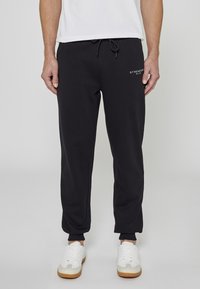 Czarne joggery wykonane z miękkiego materiału, z elastycznym paskiem z troczkiem, ściągaczami przy kostkach oraz białym logo.