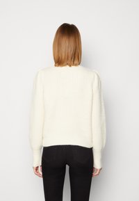 Maglione bianco lavorato a maglia con orlo a coste e motivo testurizzato. Presenta maniche a sbuffo e scollo rotondo, abbinato a jeans scuri.