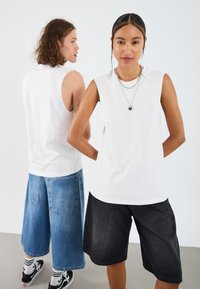 Dva modeli nosijo brezrokavne bele topi, moški model v modrih jeans kratkih hlačah in ženski model v črnih kratkih hlačah, oba z različnimi dodatki.