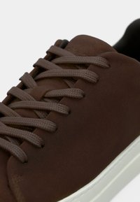 Sneakers in pelle marrone con lacci piatti marroni, texture liscia, suola in gomma bianca e design minimalista. Focus su comfort e durata.