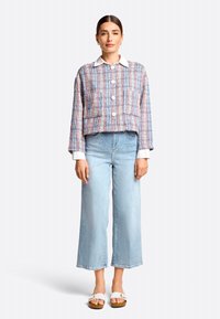 Mehrfarbige Tweedjacke mit weißem Kragen, zwei Fronttaschen und Knopfverschluss, kombiniert mit hellblauen weit geschnittenen Jeans und weißen Sandalen.