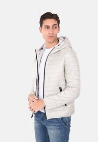 Giacca trapuntata grigia chiara con cappuccio, dotata di zip frontale, accenti neri e due tasche laterali con zip. Indossata sopra una maglietta bianca.