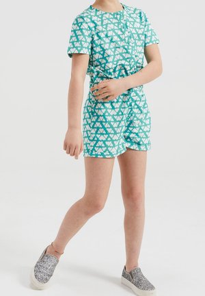 Combinaison en tissu turquoise à motifs de cœurs blancs, manches courtes, et taille élastique. Associée à des chaussures à enfiler scintillantes.