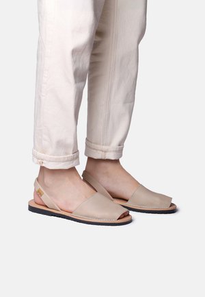 Pies con sandalias slingback de cuero beige con punta abierta combinadas con pantalones blancos sin brillo remangados sobre un fondo claro.