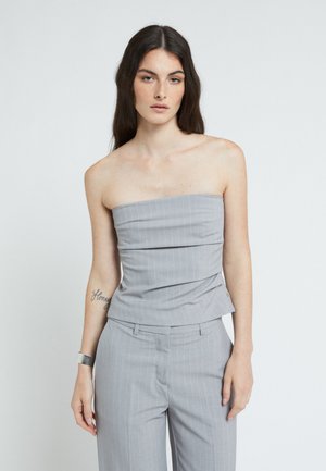 Mujer con cabello oscuro y largo, vestida con un top gris sin tirantes y pantalones a juego, de pie frente a un fondo blanco.
