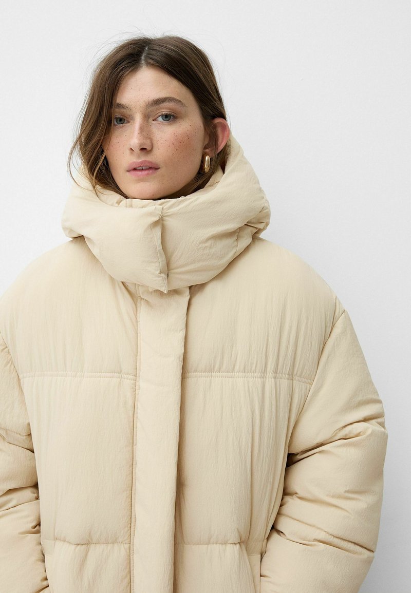 Giacca a vento beige oversize con alto colletto, caratterizzata da sezioni trapuntate e una texture liscia, adatta per climi freddi.