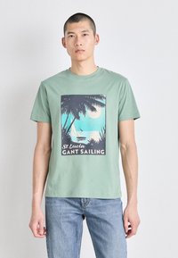 Zelené bavlněné tričko s krátkými rukávy, na kterém je grafický potisk palem, vody a měsíce. Text zní "St Lucia GANT Sailing."