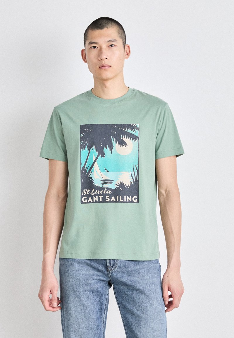 Zelené bavlněné tričko s krátkými rukávy, na kterém je grafický potisk palem, vody a měsíce. Text zní "St Lucia GANT Sailing."