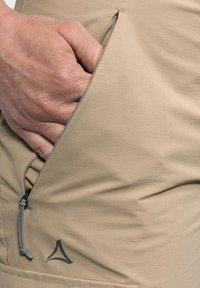 Beige Hosen mit einer Reißverschlusstasche, ausgestattet mit einem gerafften Kordelzug und einem grauen Logo. Der Stoff hat ein glattes, leichtes Aussehen und eine angenehm weiche Textur.