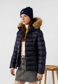 Giacca imbottita blu navy con cappuccio foderato in pelliccia, caratterizzata da un design trapuntato e dettagli con cerniere rosse. Indossata sopra un dolcevita chiaro e una gonna grigia.