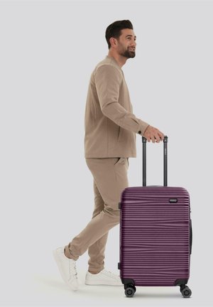 Man in een beige outfit die loopt met een paarse harde koffer met vier wielen en een uitschuifbare handgreep tegen een grijze achtergrond.