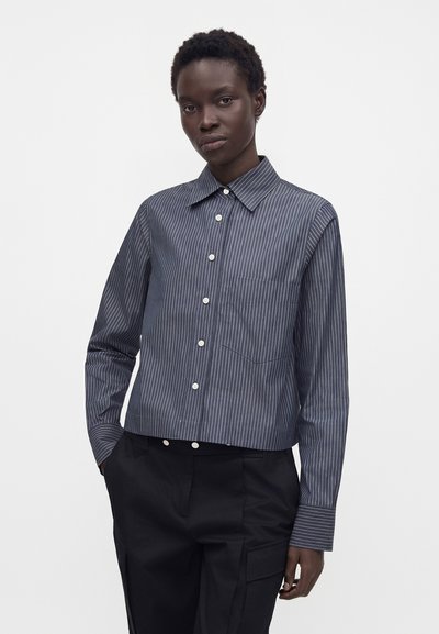 Personne portant une chemise bleu marine à fines rayures boutonnée avec des manches longues et un pantalon noir, debout avec une main dans la poche.