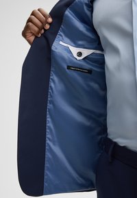 Hand, die ein offenes, marineblaues Blazer hält, das mit blauer Satinfütterung und einer geknöpften Innentasche versehen ist, die mit "Finest Stretch Fabric" beschriftet ist.
