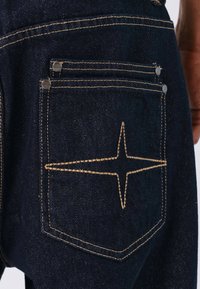 Detalle de jeans de mezclilla oscura que muestra un bolsillo trasero con costuras visibles en color naranja y herrajes plateados, presentando un diseño en forma de estrella en las costuras.