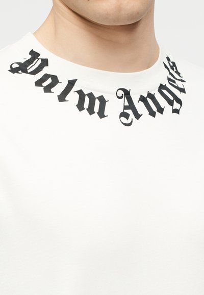 Κοντινό πλάνο λευκού πουλόβερ με λαιμόκοψη τύπου crewneck, με την επιγραφή "Palm Angels" σε μαύρη γοτθική γραμματοσειρά γύρω από το λαιμό.