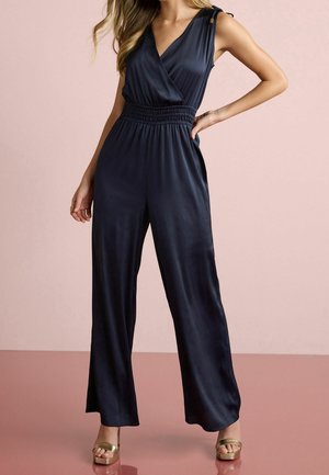 Niebieski kombinezon z dekoltem w kształcie litery V, zapinany na zakładkę, z elastycznym pasem i szerokimi nogawkami. Tkanina ma gładką, błyszczącą teksturę. Akcenty z złotego metalu.