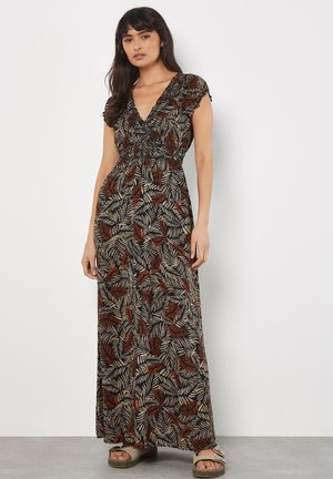 Femme portant une robe maxi noire avec des motifs de feuilles rouges et beiges, manches courtes, col en V, et sandales beiges, debout contre un mur blanc.