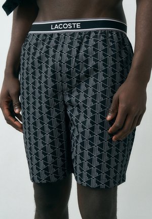 Vyriškas torsas dėvi juodus šortus su balta geometrine rašto ir juodu juosmens diržu su užrašu „LACOSTE“, stovintis priešais paprastą foną.