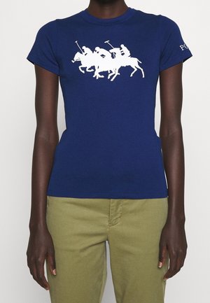 Kvinde iført en tætsiddende marineblå t-shirt med motiv af tre heste-polo-spillere i hvid, kombineret med olivengrønne bukser.
