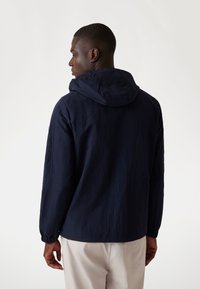 Marineblauwe vest met capuchon, met een gestructureerde afwerking, elastische boorden en een verstelbare capuchon, gezien van achteren op een lichte achtergrond.