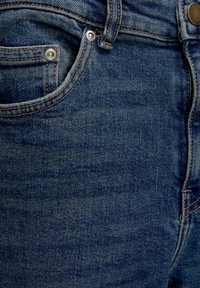 Close-up of blue denim jeans focusing on front pocket, metal rivets, stitching, and waistband with button closure.