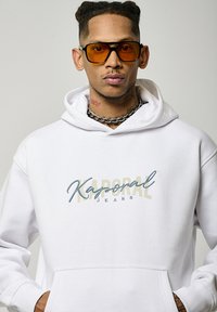 Sweat à capuche blanc avec le logo "Kaporal Jeans" en bleu et doré, doté d'une poche avant et d'une capuche à cordon. Le modèle porte des lunettes teintées orange.