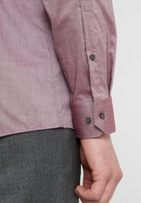 Camisa de manga larga en color burdeos claro con tejido texturizado. Presenta botones negros en el puño y un dobladillo deshilachado a lo largo del lateral.