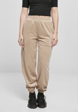 Person wearing beige velour jogger pants with elastic cuffs, black sneakers, and a black cropped top, standing against a plain background.