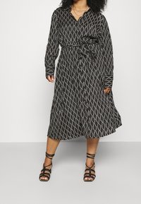 Vestido negro con estampado y cinturilla con lazo, con escote en V y mangas largas. Acentuado con un diseño geométrico blanco. Sandalias negras de tiras.