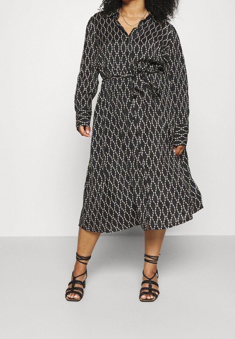 Vestido negro con estampado y cinturilla con lazo, con escote en V y mangas largas. Acentuado con un diseño geométrico blanco. Sandalias negras de tiras.