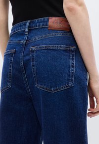 Τζιν παντελόνια σε σκούρο μπλε, με δερμάτινη ετικέτα Calvin Klein στη ζώνη, δύο πίσω τσέπες και εμφανείς ραφές.