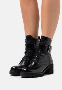 Bottes à chevilles en cuir verni noir avec un la�çage sur le devant, une sangle à boucle à la cheville et une semelle texturée épaisse. Fini brillant et détails cousus.