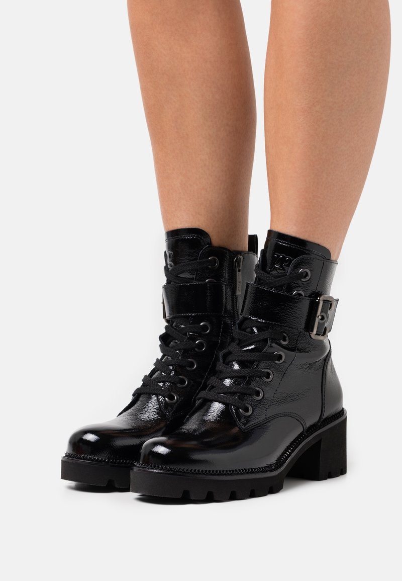 Bottes à chevilles en cuir verni noir avec un laçage sur le devant, une sangle à boucle à la cheville et une semelle texturée épaisse. Fini brillant et détails cousus.