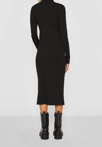 Robe noire côtelée à manches longues avec une silhouette ajustée et une longueur midi, associée à des bottines noires en cuir épaisses.