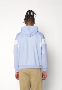 Ljusblå hoodie med kenguruficka, vita detaljer på axlarna och ribbade ärmslut. Slät tyg med avslappnad passform och justerbar huva.