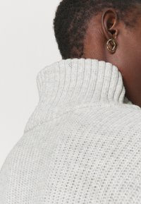 Maglione grigio a coste con colletto alto, caratterizzato da un design a maglia texturizzata. Un orecchino a cerchio dorato è visibile sull'orecchio in profilo.
