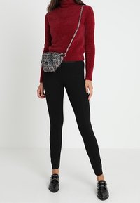 Maglione a collo alto rosso in pelliccia sintetica, leggings neri e mocassini neri con fibbie; la modella tiene in mano una piccola borsa multicolore con tracolla a catena.