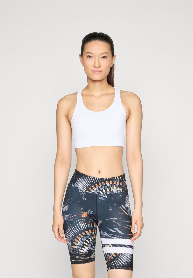 Biely športový crop top so širokými ramienkami skombinovaný s čiernymi šortkami s vysokým pásom, ktoré majú farebné abstraktné vzory a biele horizontálne pruhy.