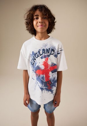 Junge mit lockigem Haar, der ein weißes T-Shirt mit England-Fußballgrafik und Jeans-Shorts vor einem einfarbigen Hintergrund trägt.