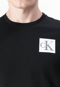 T-shirt en coton noir avec un col rond et un patch carré blanc arborant les caractères imprimés "cK" dans un design rayé.