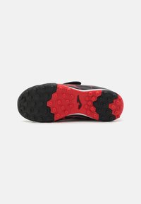 Suela de zapatilla deportiva en negro y rojo con un patrón hexagonal, que presenta goma negra y acentos rojos. Se observa una correa de velcro en la parte superior.